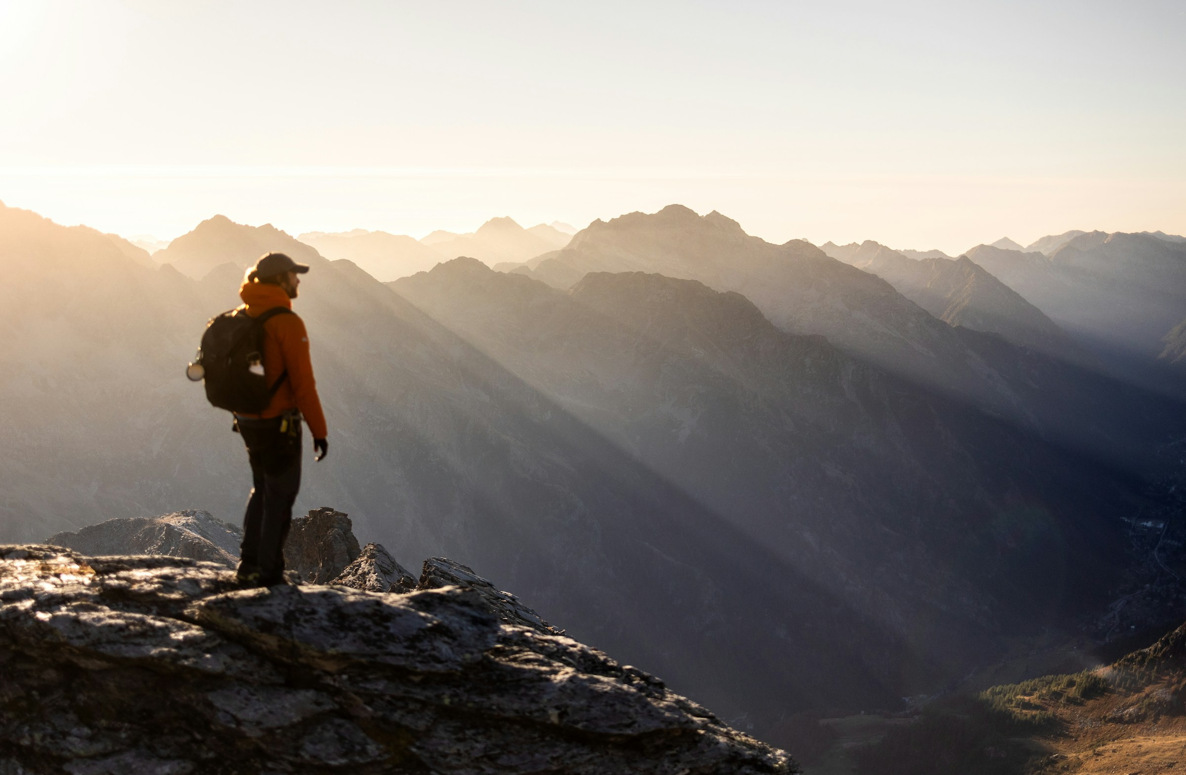 A hiker with a backpack standing on a rocky mountain peak, looking out over a sunlit mountain range.